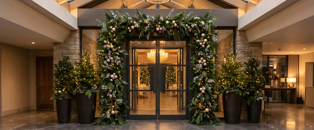 Hotel entrance framed with lit evergreen garland and gold ornaments, flanked by holiday trees.