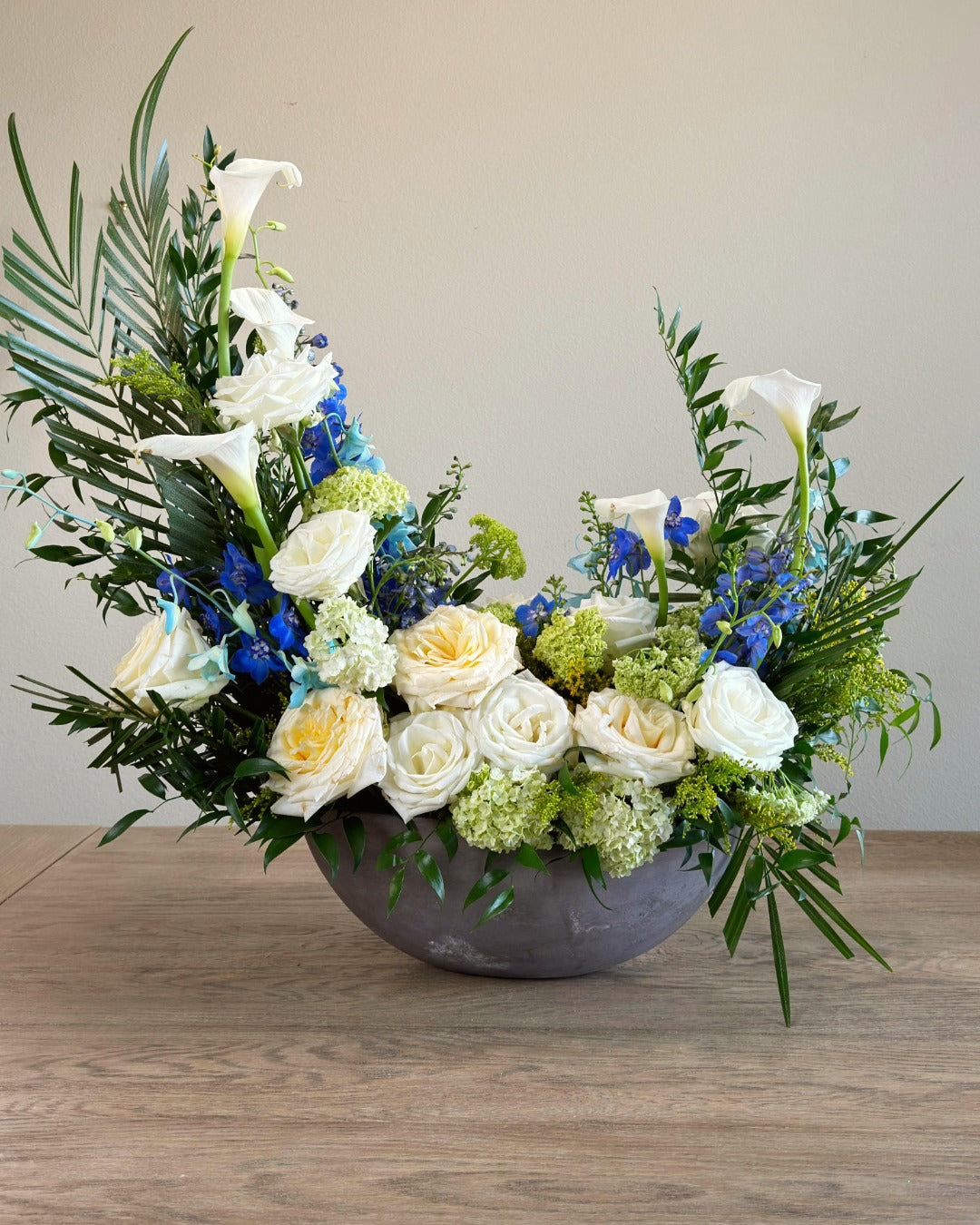 Floral arrangement with white, yellow, and blue flowers in a gray bowl on a wooden surface.