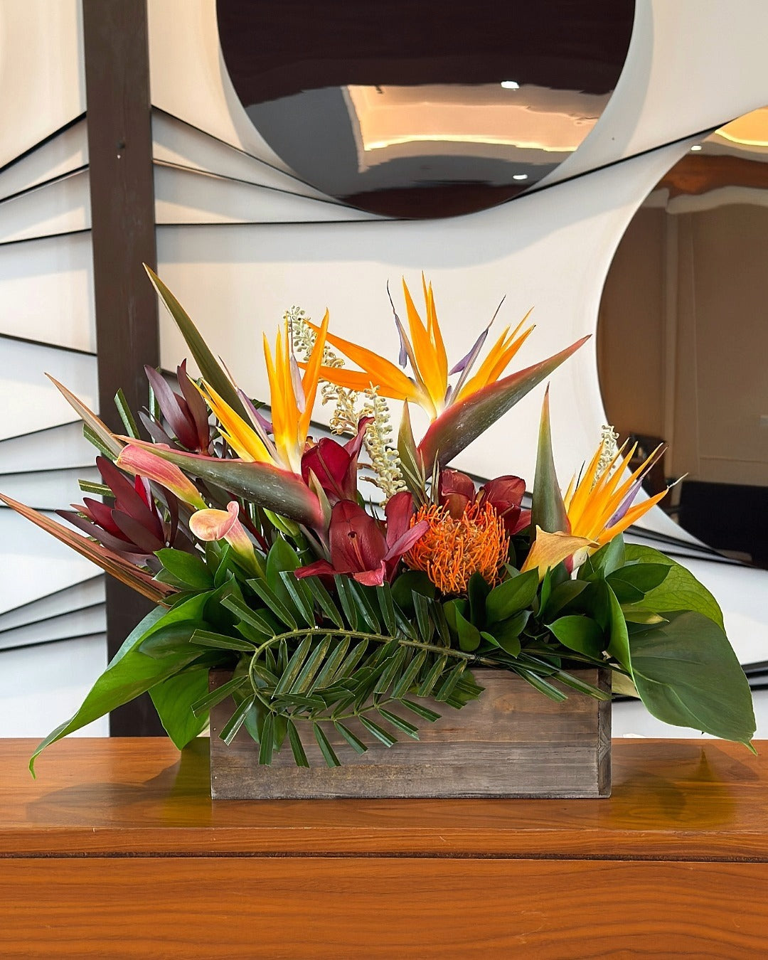 A tropical hotel reception arrangement featuring bird of paradise, deep burgundy lilies, safari sunset, and tropical leaves.
