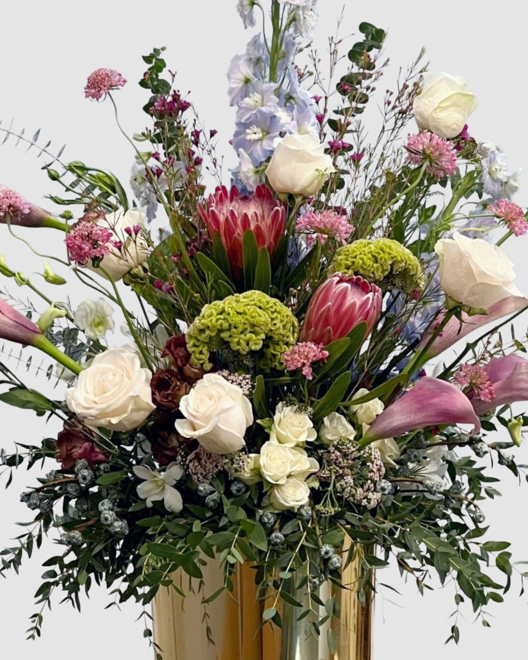 Luxury premium floral arrangement with protea, roses, delphinium, calla lilies, and textural garden blooms in a gold vessel.