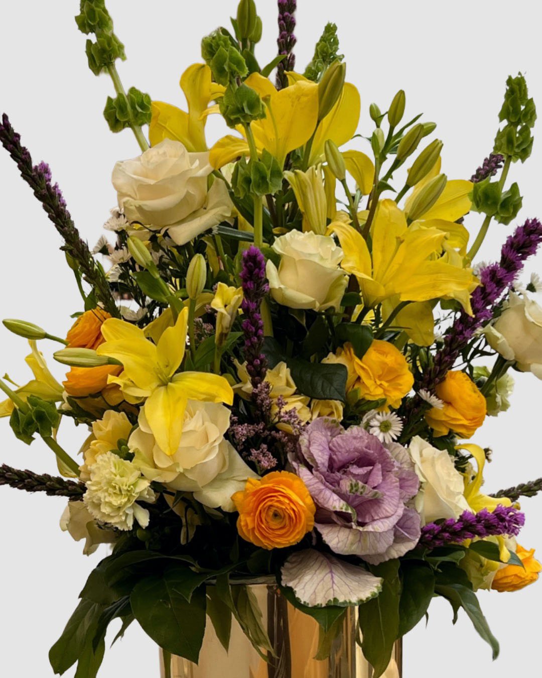 Garden-style floral arrangement in yellow and purple accents with lilies, cabbage roses, ranunculus, and bells of Ireland.