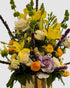 Garden-style floral arrangement in yellow and purple accents with lilies, cabbage roses, ranunculus, and bells of Ireland.