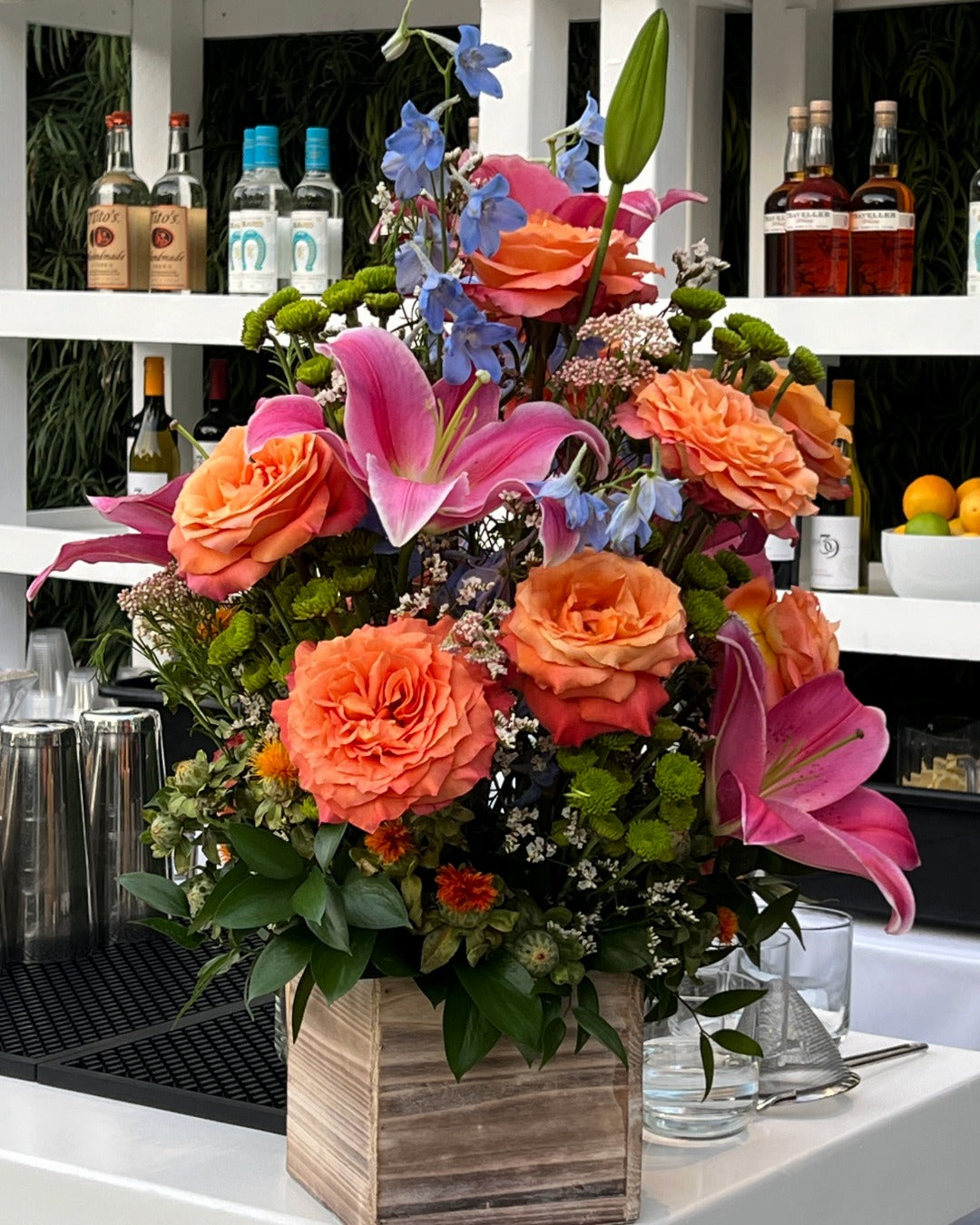 Bar floral arrangement with sunset roses, baby blue delphinium, and hot pink lilies in a wooden box container.