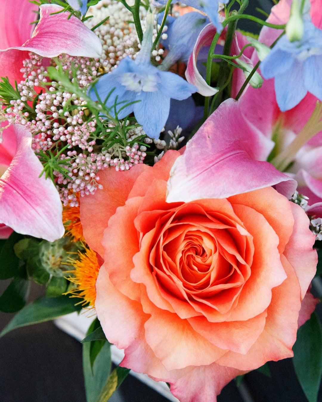 Closeup of event florals with sunset roses, hot pink lilies, baby blue delphinium, and rice flower.