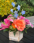 Floral arrangement with pink, orange, and blue flowers in a wooden box on a dark surface.