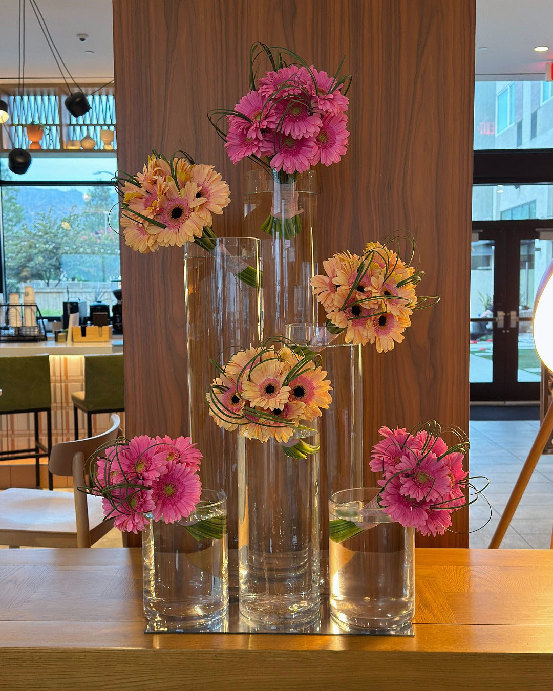 Hotel lobby floral installation with six hot pink and peach gerbera daisy bouquets forming interlocking triangles in glass cylinder vases.