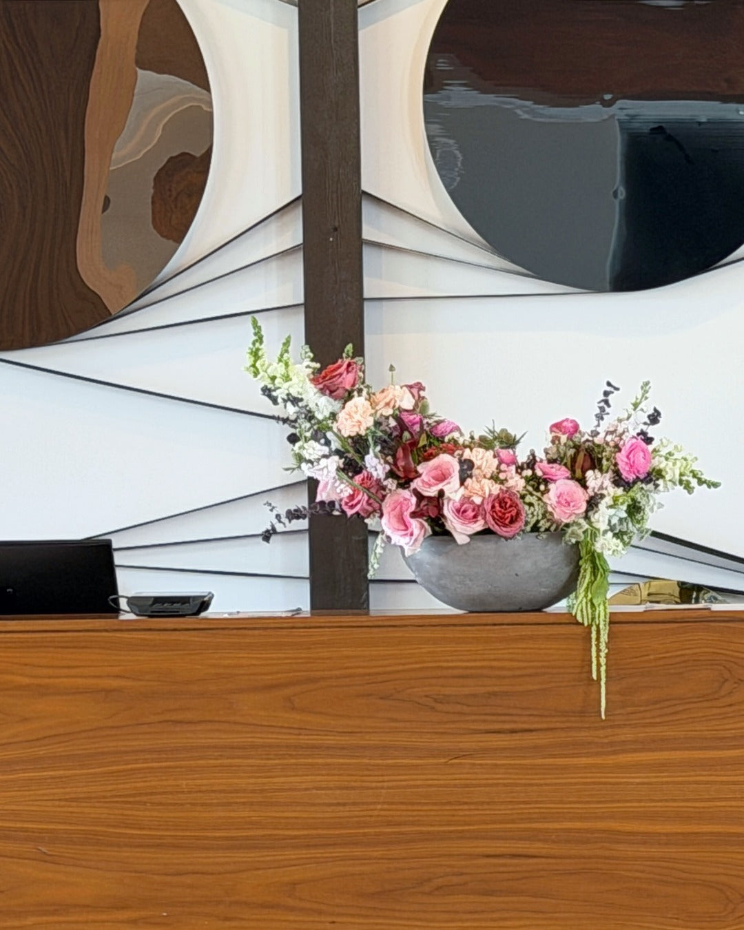 Hotel lobby check-in desk floral arrangement in a concrete vase on a wooden surface with abstract art in the background.