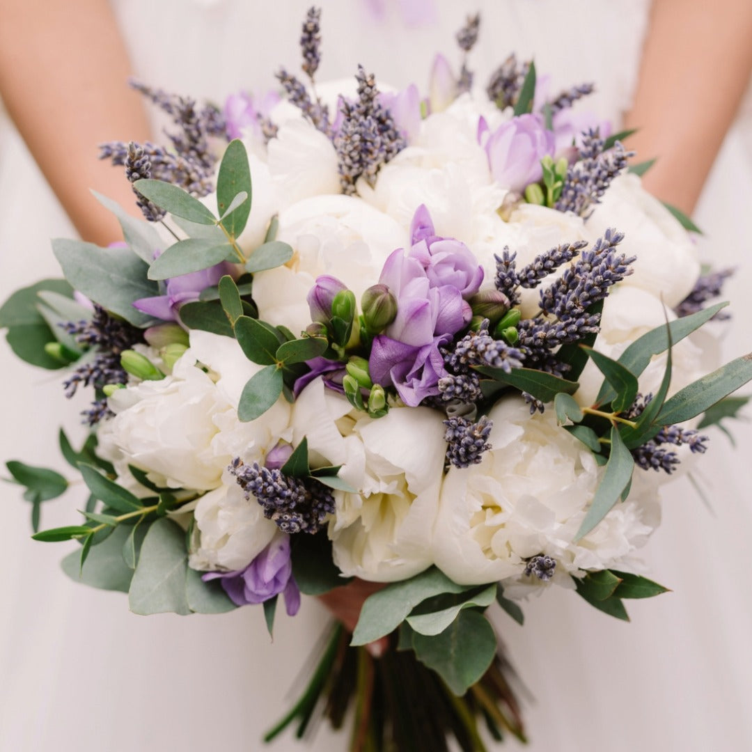 A close-up of a bridal bouquet featuring white peonies, purple anemones, lavender sprigs, and green eucalyptus leaves.