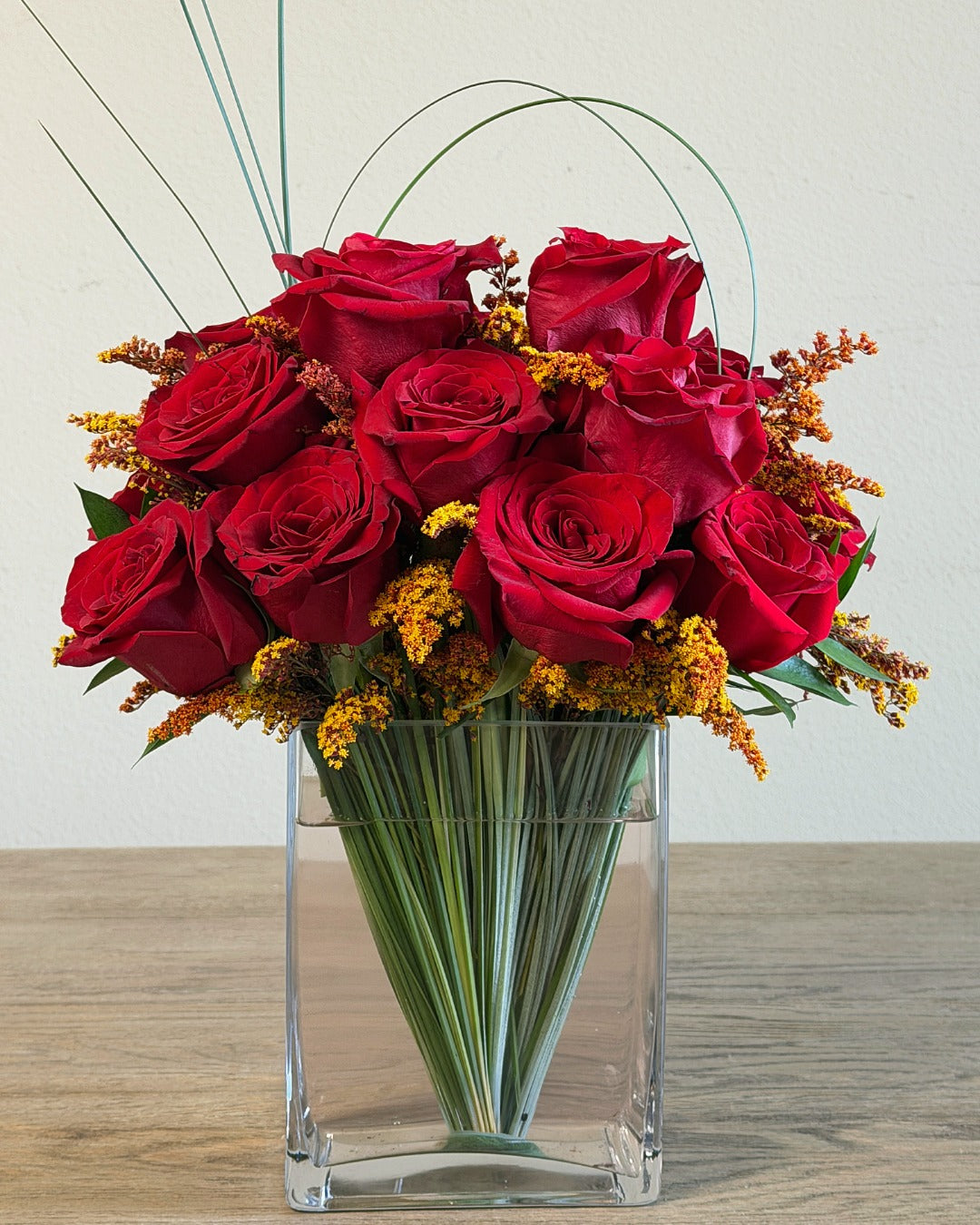 A bouquet of 18 red roses with orange-tinted aster solidago arranged in a clear glass vase with bear grass concealing the stems.