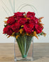 360-degree arrangement of red roses and orange-tinted solidago in a rectangular glass vase with sculptural bear grass.