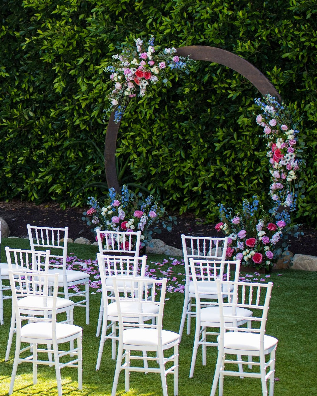 Garden-style floral arch with rose petals on the ground  and white guest chairs for an outdoor vow renewal ceremony