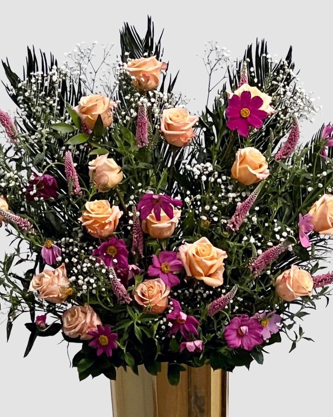 Lush garden-style arrangement with roses, cosmos, veronica, and baby’s breath in a gold vessel.