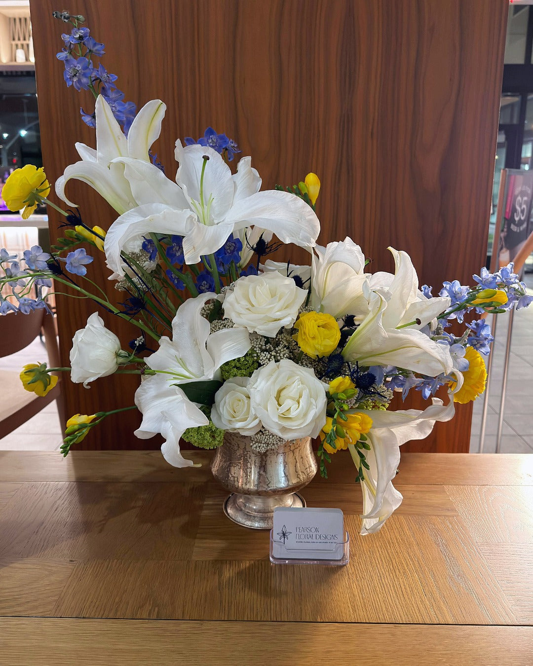 Floral arrangement with white lilies and yellow roses in a silver vase on a wooden surface.