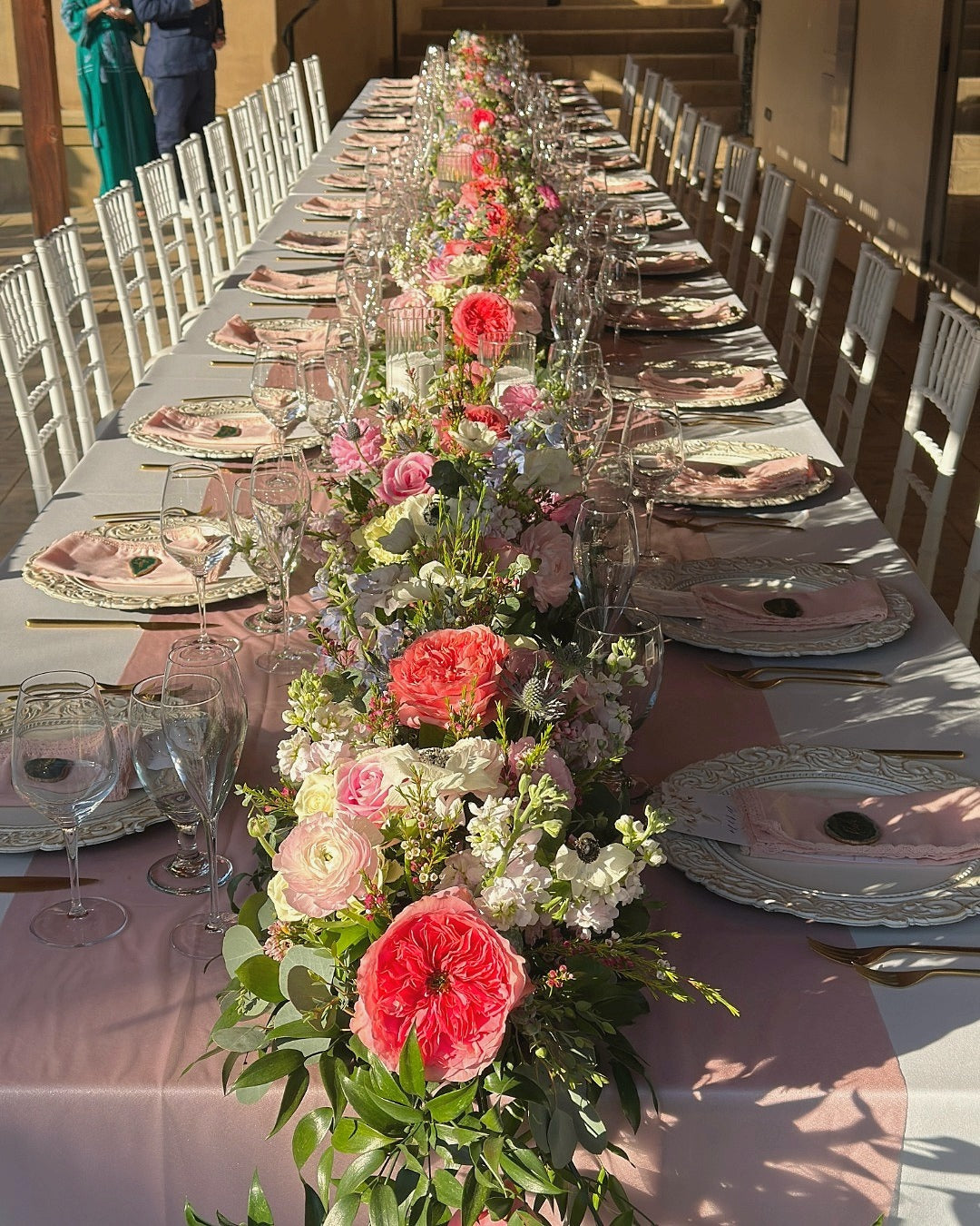 Long table set for a meal with floral centerpieces, plates, and glasses outdoors.
