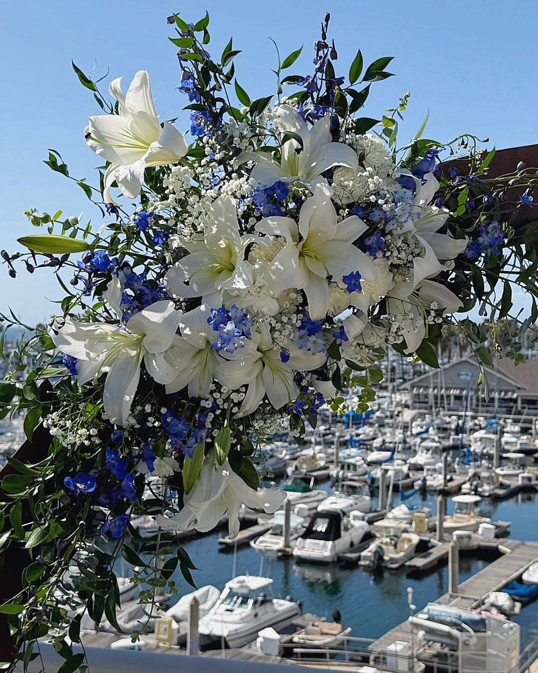 Wedding arch close-up of top installation featuring lilies, delphinium, carnations, baby's breath and greens against a marina background.